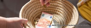 Geflochtener Korb mit Geldscheinen und Münzen darin. Eine Hand hält den Korb eine andere wirft eine Münze hinein.