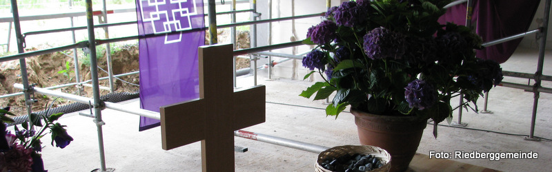 Baustelle - Baugerüste im Hingergrund - davor ein improvisierter Altar mit Holzkreuz und Blumen. Das Facettenkreuz (weiß auf lila) als Fahne