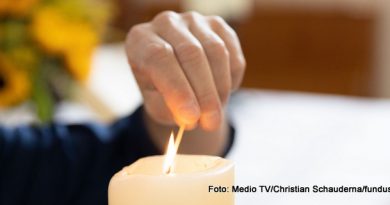 Eine Hand kommt von links ins Bild und zündet die weiße Kerze in der Bildmitte an.
