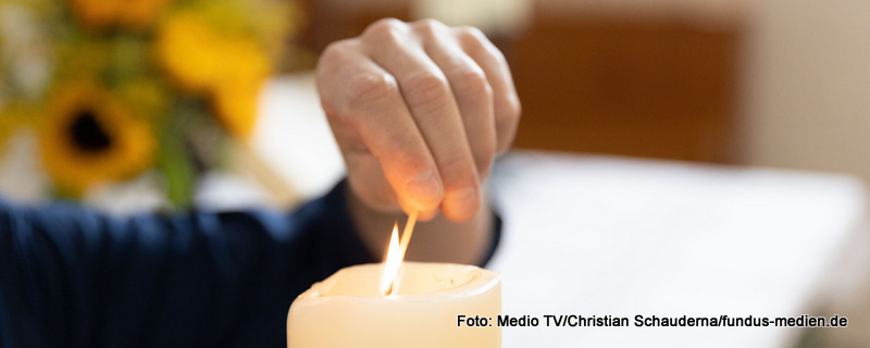 Eine Hand kommt von links ins Bild und zündet die weiße Kerze in der Bildmitte an.