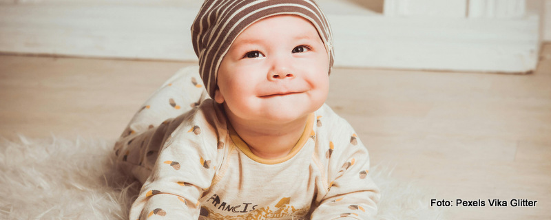 Ein Baby liegt auf dem Boden - stützt sich mit den Händen ab und lächelt in die Kamera. Es ist beigefarben gekleidet und trägt eine Mütze.
