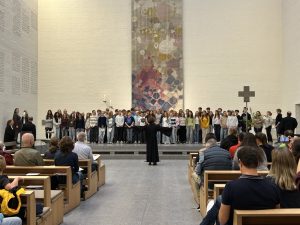 Eine große Gruppe Jugendlicher steht in einem weiten Kirchenraum - auf der rechten Seite steht ein Kreuz - vor der Gruppe steht eine Pfarrerin mit ausgebreiteten Armen zum Segen der Jugendlichen. In den Kirchenbänken sitzen zahlreiche Besucherinnen und Besucher.