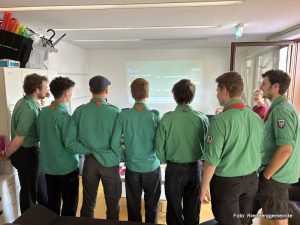Pfadfinder mit grünen Hemden stehen vor einer Karaoke-Wand und singen.