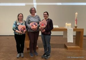Drei Frauen mit Blumen in den Händen stehen vor dem Altar der Riedberggemeinde, die Kerzen auf dem Altar brennen, das Lichtkreuz an der Altarwand leuchtet.
